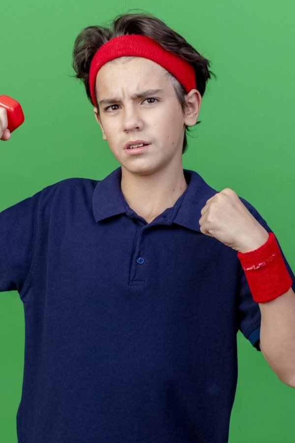 confident young handsome sporty boy wearing headband and wristbands with dental braces raising up dumbbell clenching fist looking at camera isolated on green background with copy space Does Working Out Really Stop Height Growth